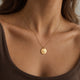 Model in a mocha-brown top wearing a personalized round gold necklace engraved with text and a date, with space for engraving of text, coordinates or handwriting on the front and back