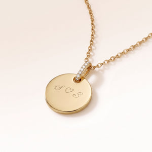 Close-up of a round gold pendant with a zirconia-set bail and personalised engraving of initials with a heart
