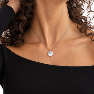 Model in a black top wearing a round silver necklace with a ridged sunburst pattern and room for personal engraving of text, coordinates or handwriting on the back